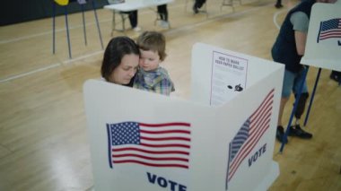 Elinde bebek olan kadın seçmen oy kabininde duruyor. Amerikan vatandaşları oy kullanma merkezine oy vermeye geldi. ABD başkan adaylarının siyasi ırkları. Ulusal Seçim Günü. Vatandaşlık görevi. Dolly vurdu..