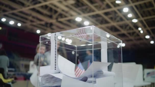 ポーリングステーションのテーブルの上に立っている投票箱の近くのショット 子供の手を握ったアメリカの女性有権者は 掲示板を箱に入れる アメリカ合衆国大統領候補の政治的競争 — ストック動画