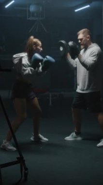 Boks eldivenli kadın boksörün dikey vuruşu, boks salonunda koçla yumruk atma ve dövüş teknikleri üzerinde pratik yapma vuruşuna denk geliyor. Atletik kadın spor yapıyor ve maç için hazırlanıyor..