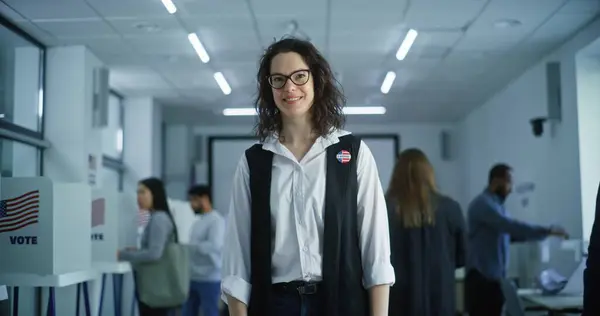 stock image Woman with badge stands in polling station, poses, smiles, looks at camera. Portrait of Caucasian woman, United States of America elections voter. Background with voting booths. Concept of civic duty.