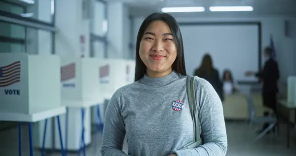 stock image Portrait of Asian woman, United States of America elections voter. Woman with badge stands in modern polling station, poses, smiles, looks at camera. Background with voting booths. Civic duty concept.