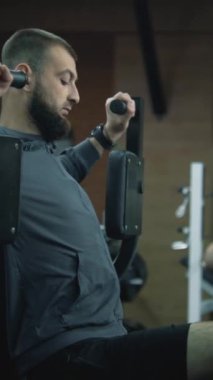 Modern fitness merkezinde spor aletleri ile egzersiz makinesinde protez bacaklı bir adamın dikey görüntüsü. Arka planda başka bir yetişkin sporcu kol egzersizi yapıyor..