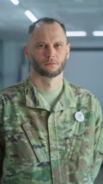 Kamuflaj üniformalı bir adamın dikey görüntüsü modern bir seçim merkezinde kameraya bakıyor. Erkek askerin portresi, Amerika Birleşik Devletleri seçmeni. Oy kullanma kabinleri olan arkaplan