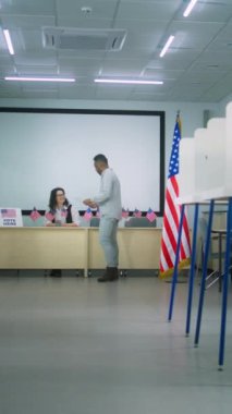 Afro-Amerikalı adamın dikey çekimi oy kullanma merkezindeki kadın oy pusulasından oy pusulasına gidiyor ve oy kabinine gidiyor. Amerika Birleşik Devletleri 'nde Ulusal Seçim Günü. Vatandaşlık görevi