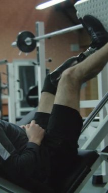 Modern fitness merkezinde bacak pres makinesi üzerinde protez bacak egzersizi yapan dikey şutçu adam. Fiziksel engelli motive bir sporcu profesyonel spor ekipmanları kullanarak güçlü bir antrenman yapıyor.