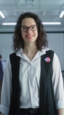 Rozetli bir kadının dikey görüntüsü sandık merkezinde duruyor, poz veriyor, gülümsüyor, kameraya bakıyor. Beyaz kadın portresi, Amerika Birleşik Devletleri seçmeni. Oy kullanma kabinleri olan arkaplan. 