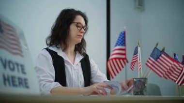 Kadın anket memuru oy kullanma merkezindeki Amerikan bayraklarıyla masada oturuyor ve oy kaydı hakkında konuşuyor. Birleşik Devletler 'de Ulusal Seçim Günü. Vatandaşlık görevi ve demokrasi.