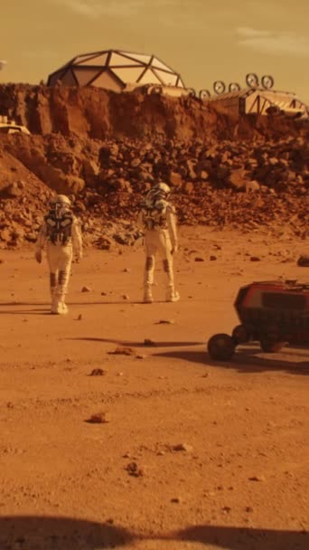 两名身穿宇航服的宇航员在火星表面行走 研究站或科学基地 探索红色星球上的太空任务 未来的技术 垂直射击 — 图库视频影像