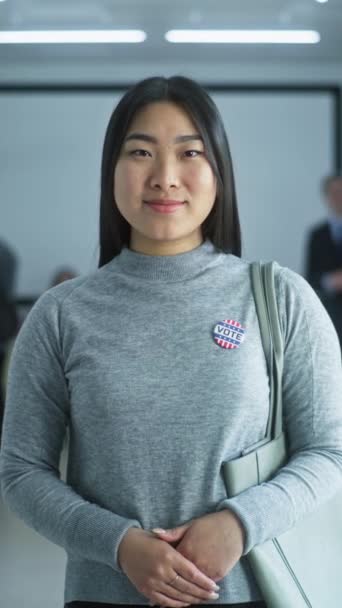 对亚洲妇女的垂直射击 美利坚合众国的选举人 拿着徽章的女人站在现代投票站 摆出姿势 看着镜头 有投票亭的背景 公民义务 — 图库视频影像