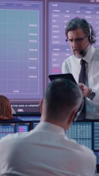 中年留着胡子的男人带着耳机 一边用平板电脑一边聊天 一边在股票交易所的专业交易员团队中工作 垂直射击 — 图库视频影像