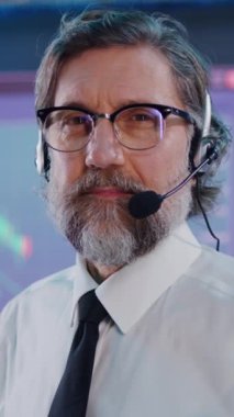 Sakallı, orta yaşlı, kulaklıklı ve gözlüklü bir adam borsada çalışırken kameraya bakıyor. Dikey çekim