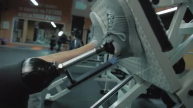 Modern spor salonunda bacak pres makinesinde protez bacak eğitimi alan motive olmuş bir sporcunun fotoğrafı. Yapay bacaklı yetişkin sporcu profesyonel spor malzemelerini kullanarak egzersiz yapıyor..