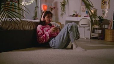 Moğol kız kulaklıkla müzik dinliyor ve rahat yatak odasında yerde oturan cep telefonuyla sosyal ağlarda sörf yapıyor. Genç kız evde eğleniyor ve eğleniyor. Yaşam tarzı kavramı.