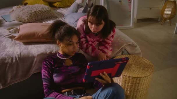モンゴル人のティーンはベッドに横たわり 友人とのコンテンツを見ています アフリカ系アメリカ人の少女が床に座り タブレットを使ってインターネットをサーフ 複数の民族の女の子が一緒に家でレジャー時間を過ごしています 友人関係 — ストック動画