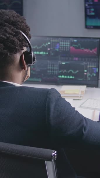 アフリカ系アメリカ人の男性トレーダーは 表示されたリアルタイム在庫でコンピュータで動作します 同僚は バックグラウンドで大きなスクリーン上の為替市場チャートを分析します 仮想通貨取引と投資 バーティカル — ストック動画