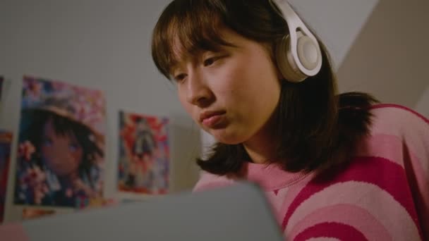 ヘッドフォンのモンゴル人の十代の少女は 彼女の居心地の良い寝室に座ってオンラインレッスンをしながら ラップトップ上のビデオ通話で話します 自宅で遠隔学習をしている学生 ライフスタイルと距離教育 — ストック動画