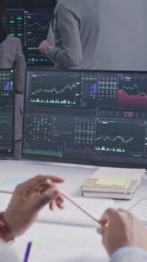 Elinde kalem olan Afrikalı Amerikalı tüccar gerçek zamanlı hisse senetleriyle bilgisayar başında çalışıyor. Meslektaşlar, arka plandaki büyük ekranlardaki döviz piyasalarının grafiklerini analiz ederler. Kripto para ticareti. Dikey