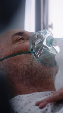 Oksijen maskeli yaşlı hasta akciğer havalandırması sırasında yatakta yatıyor. Hemşire yaşlı adamla ilgileniyor, monitör kullanıyor. Hastanedeki modern ekipmanlarla birlikte acil serviste. Dikey çekim