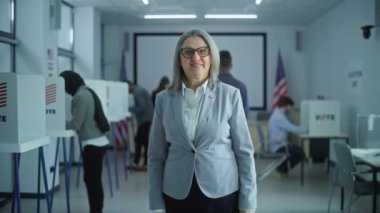 İş kadını modern bir seçim merkezinde duruyor, poz veriyor, kameraya bakıyor, gülümsüyor. Yetişkin bir kadının portresi, Amerika Birleşik Devletleri seçmeni. Oy kullanma kabinleri ve Amerikan bayrağıyla arka plan.