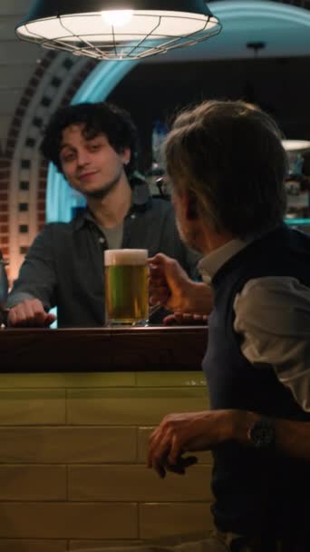 年轻酒保的垂直镜头给成熟的男性朋友们一杯啤酒 晚上两个男人坐在酒吧柜台边 在时尚的酒吧里聊天喝酒 遇见老朋友了 联合国的概念 — 图库视频影像