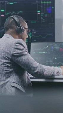 Afrikalı Amerikalı finansal analist gerçek zamanlı bilgisayar hisselerini izliyor, kalemle veri yazıyor, banka ofisinde çalışıyor. Piyasa grafikleri büyük dijital ekranlarda gösteriliyor. Yatırım ya da analiz.
