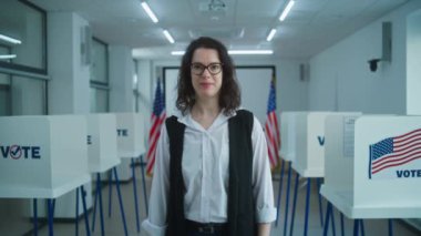 Amerikalı kadın seçmen ya da sandık memuru kameraya konuşuyor, oy kullanma çağrısı yapıyor. Birleşik Devletler 'de Ulusal Seçim Günü. Oy kullanma merkezinin arka planında oy kullanma kabinleri var. Vatandaşlık görevi ve vatanseverlik.