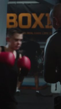 Odaklanmış genç dövüşçünün dikey vuruşu boks salonunda antrenman yaparken kum torbasına çarpıyor. Boks eldivenli genç sporcu turnuvadan önce koçla egzersiz yapıyor..