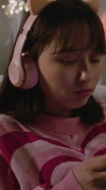 Asyalı kız kulaklıkla müzik dinliyor ve rahat yatak odasındaki halının üzerinde oturarak internette geziyor. Genç kız boş zamanlarını evde eğlenerek geçiriyor. Dikey çekim