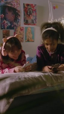 Yatak odasında çok ırklı iki genç kız, internette akıllı telefonlarıyla gezinip kulaklıkla müzik dinlerken konuşup gülüyorlar. Dikey çekim