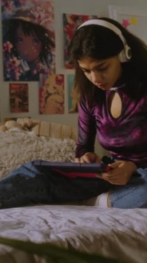Kulaklıklı güzel bir genç odasında yatağında oturuyor ve tablet kullanarak internette sörf yapıyor. Afrikalı Amerikalı kız video içeriğine bakıyor ya da film izliyor. Dikey çekim