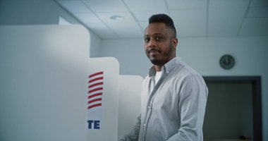 Afro-Amerikalı bir adam oy kabininde oy pusulası dolduruyor, sonra gülümsüyor ve kameraya bakıyor. Amerika Birleşik Devletleri Ulusal Seçim Günü 'nde sandık merkezinde ABD vatandaşı veya erkek seçmen.
