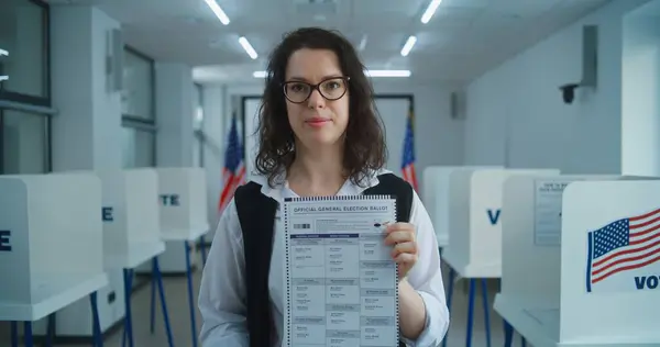 stock image American female voter or polling officer speaks on camera, shows paper ballot, calls for voting. National Election Day in the United States. Voting booths at polling station. Civic duty and patriotism