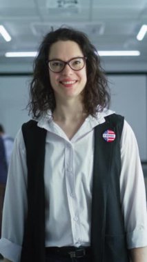 Rozetli bir kadının dikey görüntüsü sandık merkezinde duruyor, poz veriyor, gülümsüyor, kameraya bakıyor. Beyaz kadın portresi, Amerika Birleşik Devletleri seçmeni. Oy kullanma kabinleri olan arkaplan. 