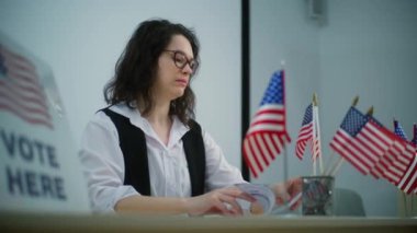 Kadın anket memuru oy kullanma merkezindeki Amerikan bayraklarıyla masada oturuyor ve oy kaydı hakkında konuşuyor. Birleşik Devletler 'de Ulusal Seçim Günü. Vatandaşlık görevi ve demokrasi.