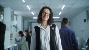 Beyaz kadın portresi, Amerika Birleşik Devletleri seçmeni. Gözlüklü kadın oy verme merkezinde duruyor, poz veriyor, gülümsüyor, kameraya bakıyor. Oy verme kabinleri olan bir arka plan. Yurttaşlık görevi kavramı.