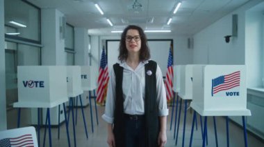 Rozetli bir Amerikan vatandaşı kameraya konuşuyor, oy verme çağrısı yapıyor. Birleşik Devletler 'de Ulusal Seçim Günü. Oy kullanma merkezinin arka planında oy kullanma kabinleri var. Vatandaşlık görevi. Dolly vurdu..