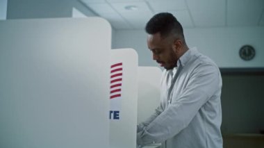 Afro-Amerikalı bir adam oy kabininde oy pusulası dolduruyor, sonra gülümsüyor ve kameraya bakıyor. Amerika Birleşik Devletleri Ulusal Seçim Günü 'nde sandık merkezinde ABD vatandaşı veya erkek seçmen.