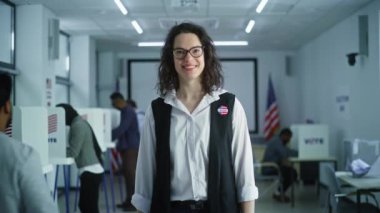 Rozetli bir kadın oy kullanma merkezinde duruyor, poz veriyor, gülümsüyor, kameraya bakıyor. Beyaz kadın portresi, Amerika Birleşik Devletleri seçmeni. Oy verme kabinleri olan bir arka plan. Yurttaşlık görevi kavramı.