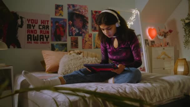 戴着耳机的美丽的非洲裔美国女孩坐在自己房间的床上 用平板电脑上网 青少年看视频内容或看电影 舒适舒适的家 室内环境舒适时尚 — 图库视频影像