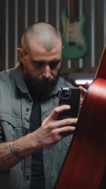 Küçük işletme sahibi, telefon kullanarak sosyal ağlar için el yapımı akustik gitarın fotoğrafını veya videosunu çekiyor. Erkek marangoz ustası, modern müzik aletleri atölyesinde çalışıyor..