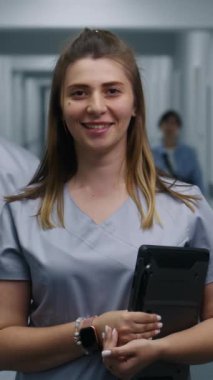 Kadın doktor modern klinik koridorunda duruyor. Profesyonel doktor dijital tableti tutuyor, gülümsüyor ve kameraya bakıyor. Hastane ya da tıp merkezi koridorundaki tıbbi personel ve hastalar.