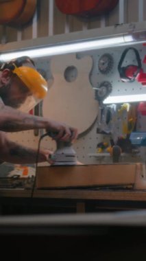 Koruyucu yüz kalkanı olan erkek marangoz, parlatma makinesi kullanarak tahta gitar gövdesini parlatır. Ahşap ustası, zanaatkar modern atölyede müzik aleti yapıyor. Dikey çekim