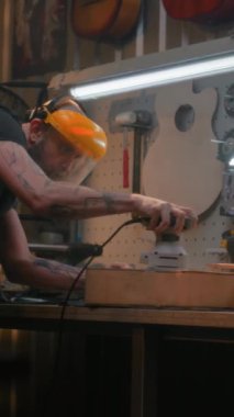 Güvenlik yüzlü erkek marangoz öğütücü kullanarak tahta gitar gövdesini öğütüyor. Ahşap ustası, zanaatkar modern atölyede müzik aleti yapıyor. El işi ve küçük işletme konsepti.