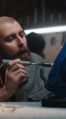 Profesyonel marangoz lehimleme demiri kullanarak gitar telleri çalar. Erkek zanaatkar modern atölyede ahşap müzik aleti yapıyor. İş arkadaşı arka planda çalışıyor. El yapımı ve girişimcilik..