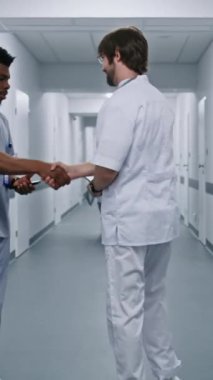 Beyaz üniformalı doktor tıp merkezi koridorunda yürüyor, Afrikalı Amerikalı meslektaşıyla selamlaşıyor ve kadın doktorla konuşuyor. Hastane koridorunda çok ırklı tıbbi personel var. Dikey çekim. Arka plan.