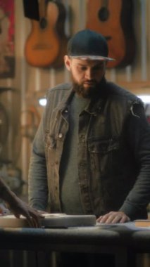 Orta yaşlı dövmeli sanatçı gitar gövdesini tutuyor ve gitar fretboard 'u deniyor. Olgun bir zanaatkar planlara bakar ve iş arkadaşlarıyla konuşur. Marangozlar şık ışık atölyesinde elektro gitar yapıyorlar..