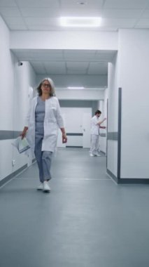 Hasta geçmişi klasörlü beyaz üniformalı doktor hastane koridorunda yürüyor. Arka planda çok ırklı sağlık uzmanları var. Tıbbi personel klinikte çalışıyor. Dikey çekim