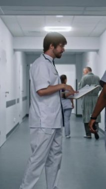 Beyaz üniformalı doktor Afrikalı Amerikalı meslektaşıyla konuşuyor ve kabineye giriyor. Doktor klinik koridorunda yürüyor. Hastanede çok ırklı tıbbi personel çalışıyor. Ağır çekim. Dikey çekim.