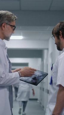 Erkek doktor MRI ve CT beyin taraması görüntülerini dijital tablet kullanan doktorlara gösteriyor. Sıhhiyeciler konuşuyor, modern klinik koridorunda durun. Hastane ya da tıp merkezi koridorundaki hasta ve sağlık personeli.