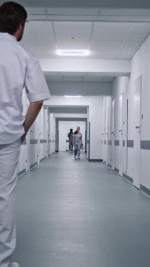 Beyaz üniformalı doktor hastane koridorunda yürür ve dolabın kapısını açmaya çalışır. Arka planda çok ırklı sağlık uzmanları var. Klinikte tıbbi personel çalışıyor. Dikey çekim. Arkadan bak. Yavaş çekim